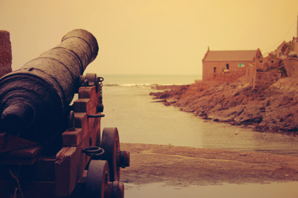 war canon pointing at ocean and a house at a cliff.